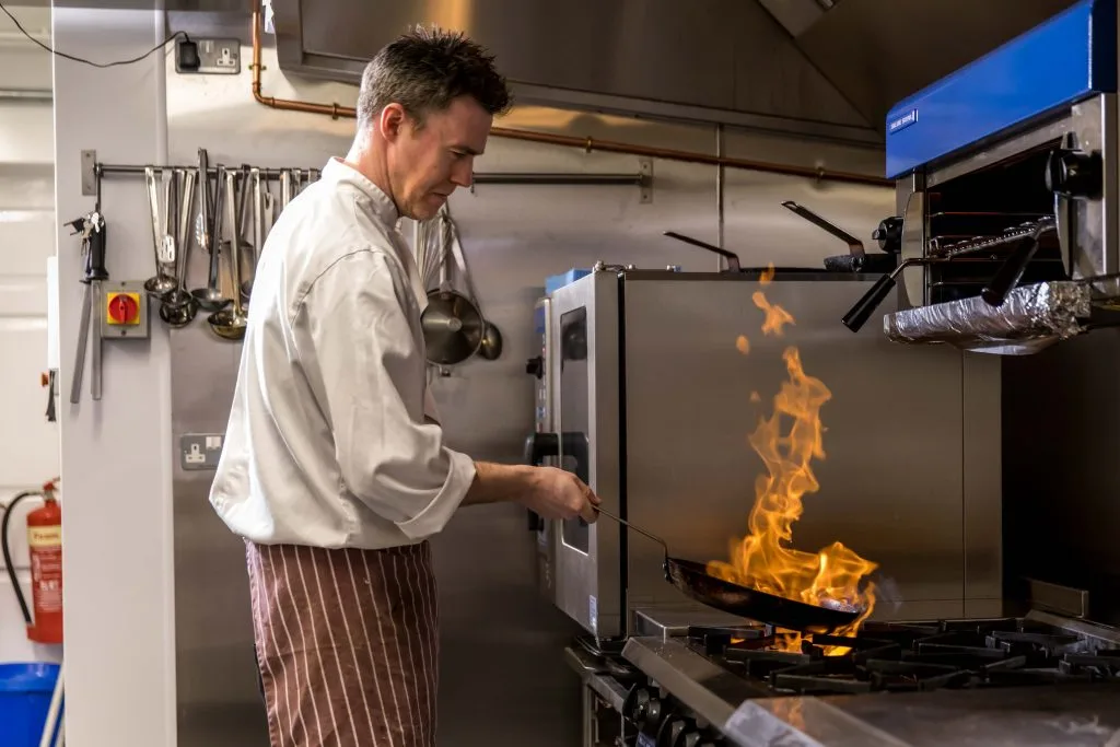 A chef holds a flaming pan above a hob in a restaurant kitchen